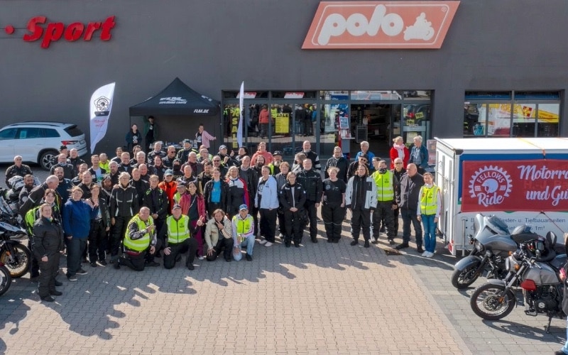 Eine große Gruppe von Teilnehmern des Fellows Ride Kiel steht für ein Gruppenfoto auf dem Parkplatz vor dem Polo-Motorradgeschäft "Motor · Freizeit · Sport" an einem sonnigen Tag. Die Fahrer tragen Motorradschutzkleidung und gelbe Warnwesten. Im Hintergrund ist das graue Gebäude mit dem "Polo"-Logo und der Aufschrift "Motor · Freizeit · Sport" zu sehen.